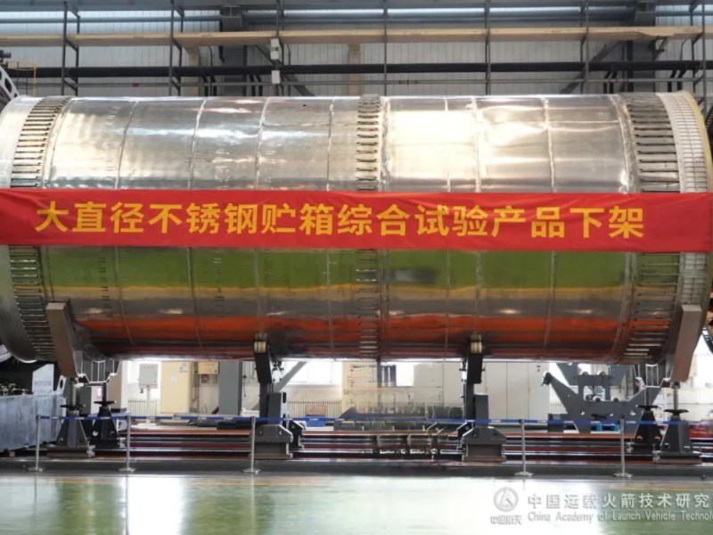 A large cylindrical stainless steel propellant tank, developed by the China Academy of Launch Vehicle Technology (CALT), sits mounted in a factory setting. A red banner across the tank reads “Large-diameter stainless steel tank integrated test product off the assembly line” in Chinese.