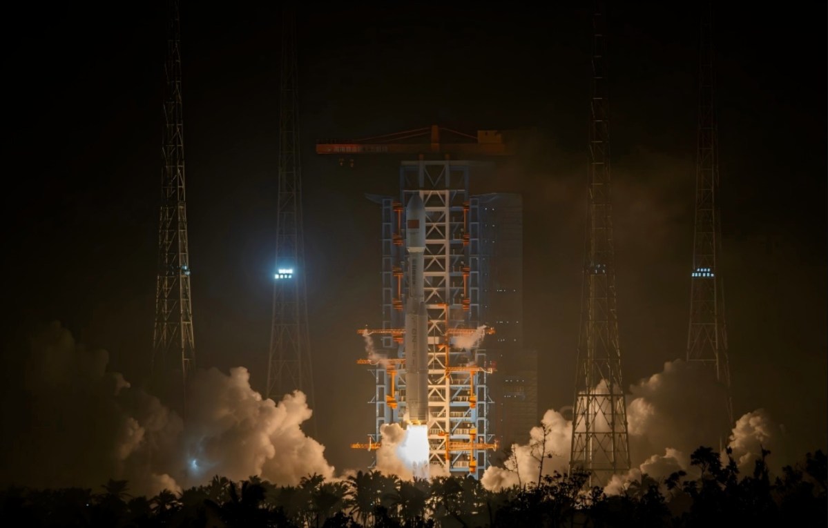 A Long March 8 rocket lifts off from Hainan Commercial Launch Site at night, illuminating the surroundings with bright flames and thick clouds of smoke. The launch pad structure and lightning towers frame the ascending vehicle against the dark sky.