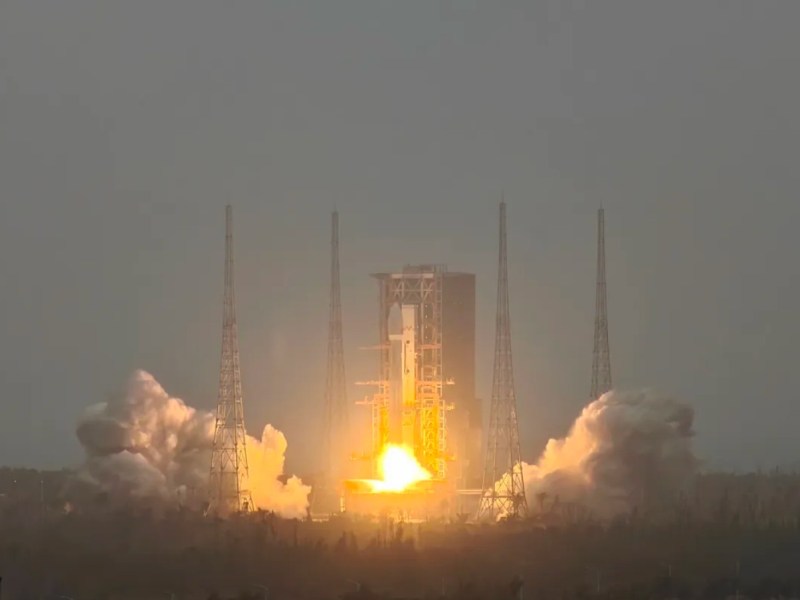A Long March 8A rocket lifts off from Wenchang Satellite Launch Center, producing bright flames and thick smoke. The rocket is surrounded by four lightning towers, with a launch pad structure partially visible in the background. The sky appears overcast, adding contrast to the fiery exhaust plume.