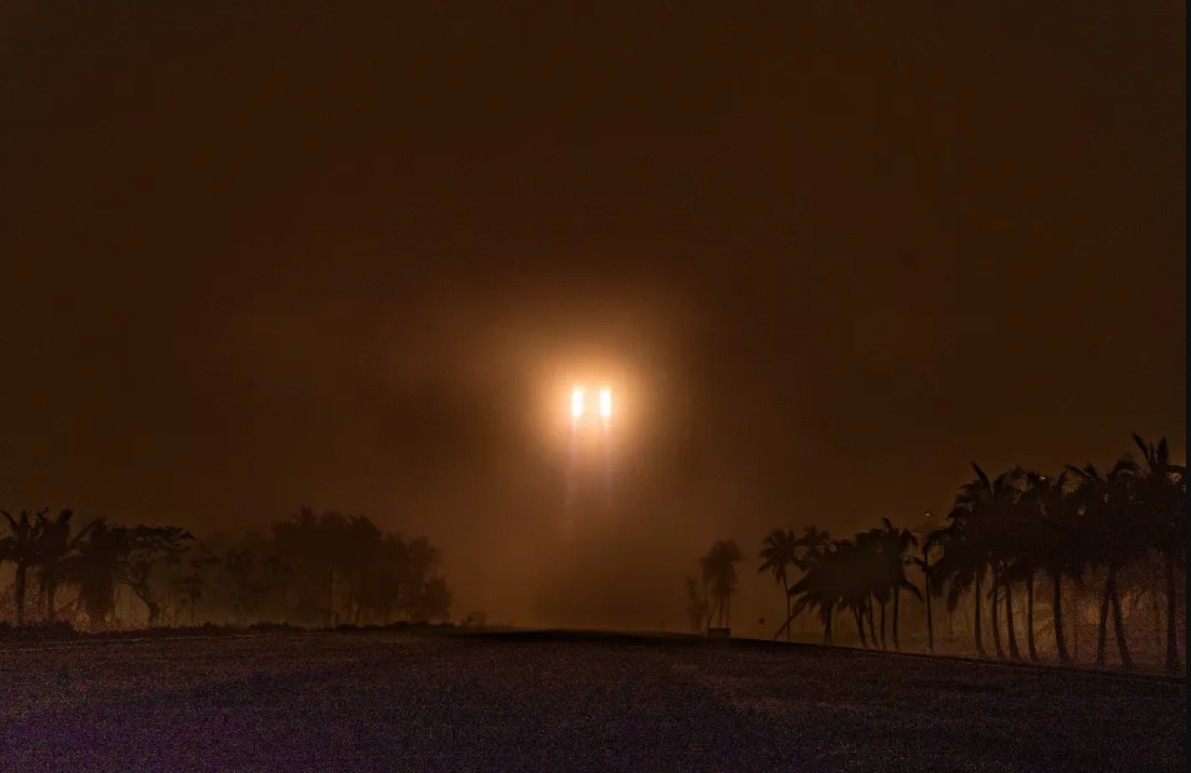 A Long March 5B rocket pierces thick fog during liftoff from Wenchang Satellite Launch Center, its bright engines casting an orange glow over silhouetted palm trees.
