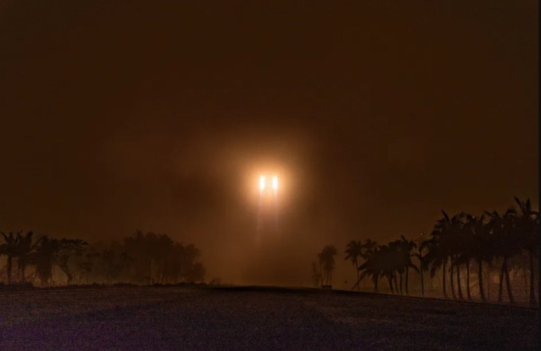 A Long March 5B rocket pierces thick fog during liftoff from Wenchang Satellite Launch Center, its bright engines casting an orange glow over silhouetted palm trees.