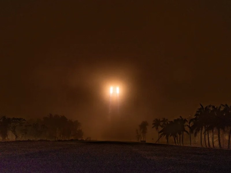 A Long March 5B rocket pierces thick fog during liftoff from Wenchang Satellite Launch Center, its bright engines casting an orange glow over silhouetted palm trees.