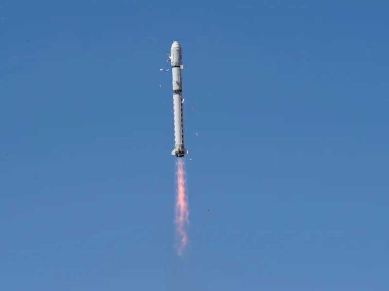 A Long March 2D rocket ascends into a clear blue sky after liftoff from Jiuquan Satellite Launch Center, trailing a pinkish-orange exhaust plume. Pieces of insulation are seen peeling away from the payload fairing during ascent.