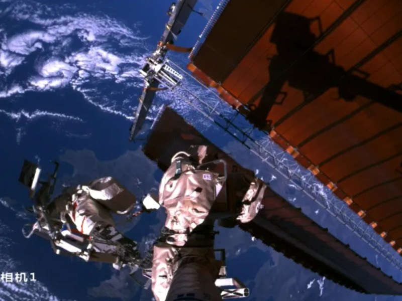 Two Chinese astronauts conducting a spacewalk outside the Tiangong space station, with Earth and clouds visible in the background. One astronaut is positioned at the end of Tiangong’s robotic arm, captured by an external camera mounted on the arm. Large solar arrays of the station are also visible.