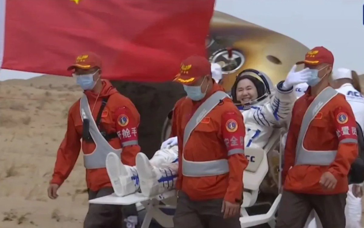 Chinese astronaut Wang Haoze smiles and waves while being carried in a reclining chair by recovery personnel after landing from the Shenzhou-19 mission, with a Chinese flag and the reentry capsule visible in the background.
