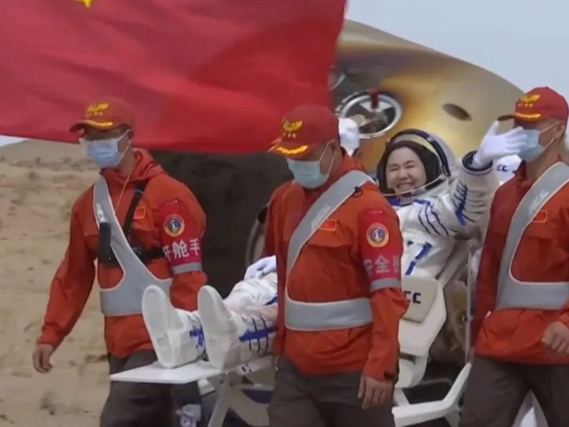 Chinese astronaut Wang Haoze smiles and waves while being carried in a reclining chair by recovery personnel after landing from the Shenzhou-19 mission, with a Chinese flag and the reentry capsule visible in the background.
