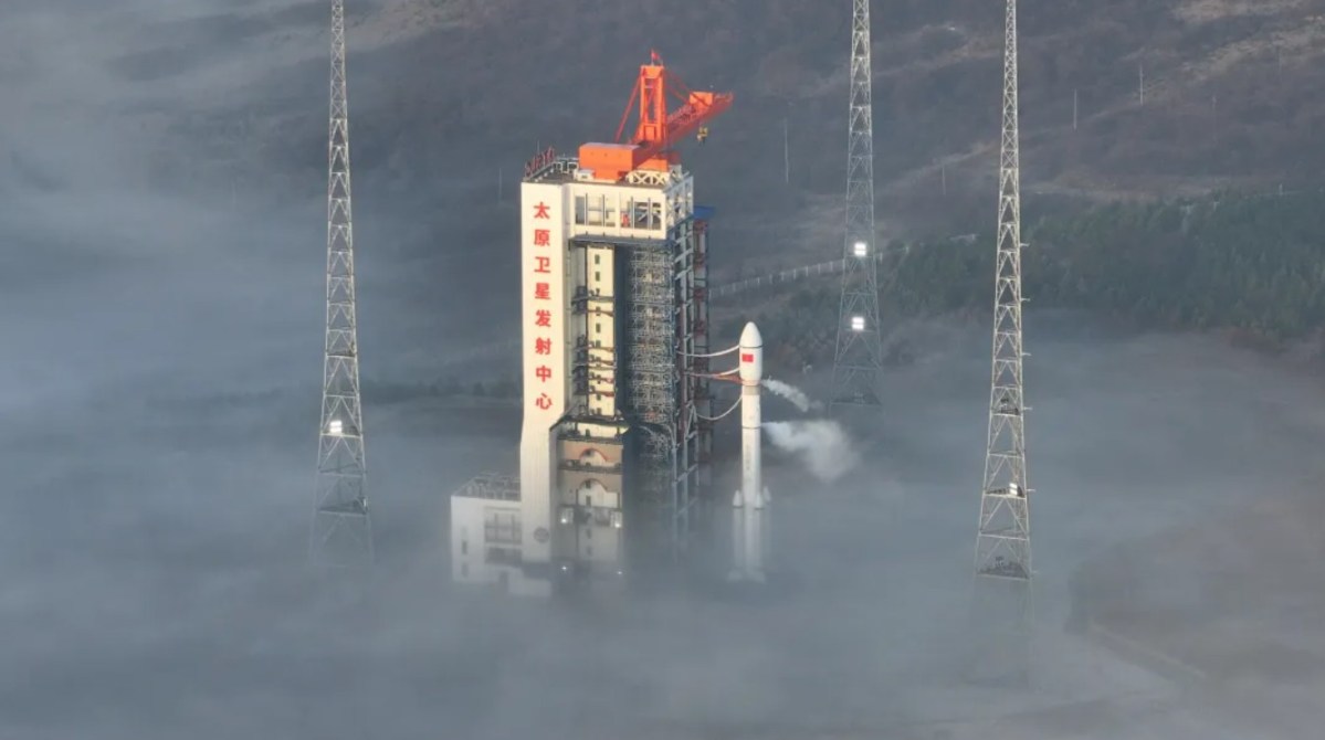 A Long March 6A rocket stands on the launch pad at China's Taiyuan Satellite Launch Center, surrounded by dense morning fog and lightning towers. Venting is visible from the rocket as it prepares for liftoff, with the launch tower displaying Chinese characters reading "Taiyuan Satellite Launch Center."